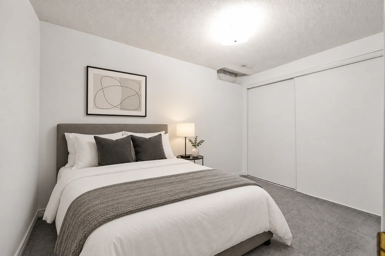 The basement bedroom virtually staged: a bed with grey bedding, a night table and lamp, a print, a closet with sliding doors, a carpeted floor