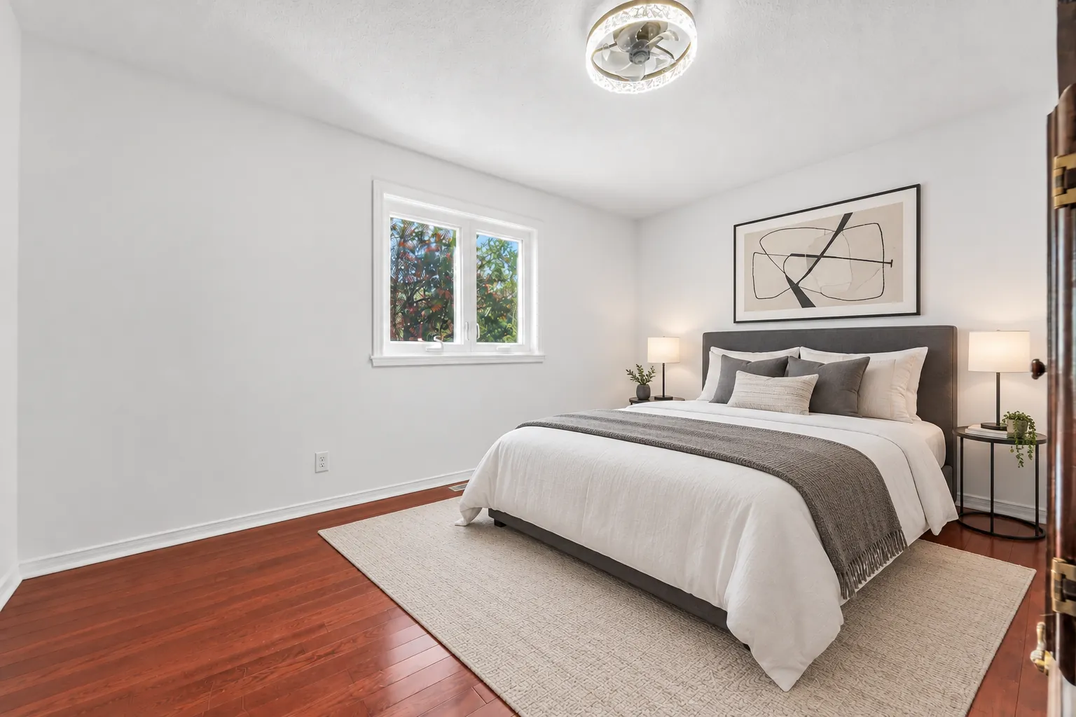 A bedroom virtually staged: an upholstered bed with grey bedding, two night tables with lamps, a rug, an abstract print, a window