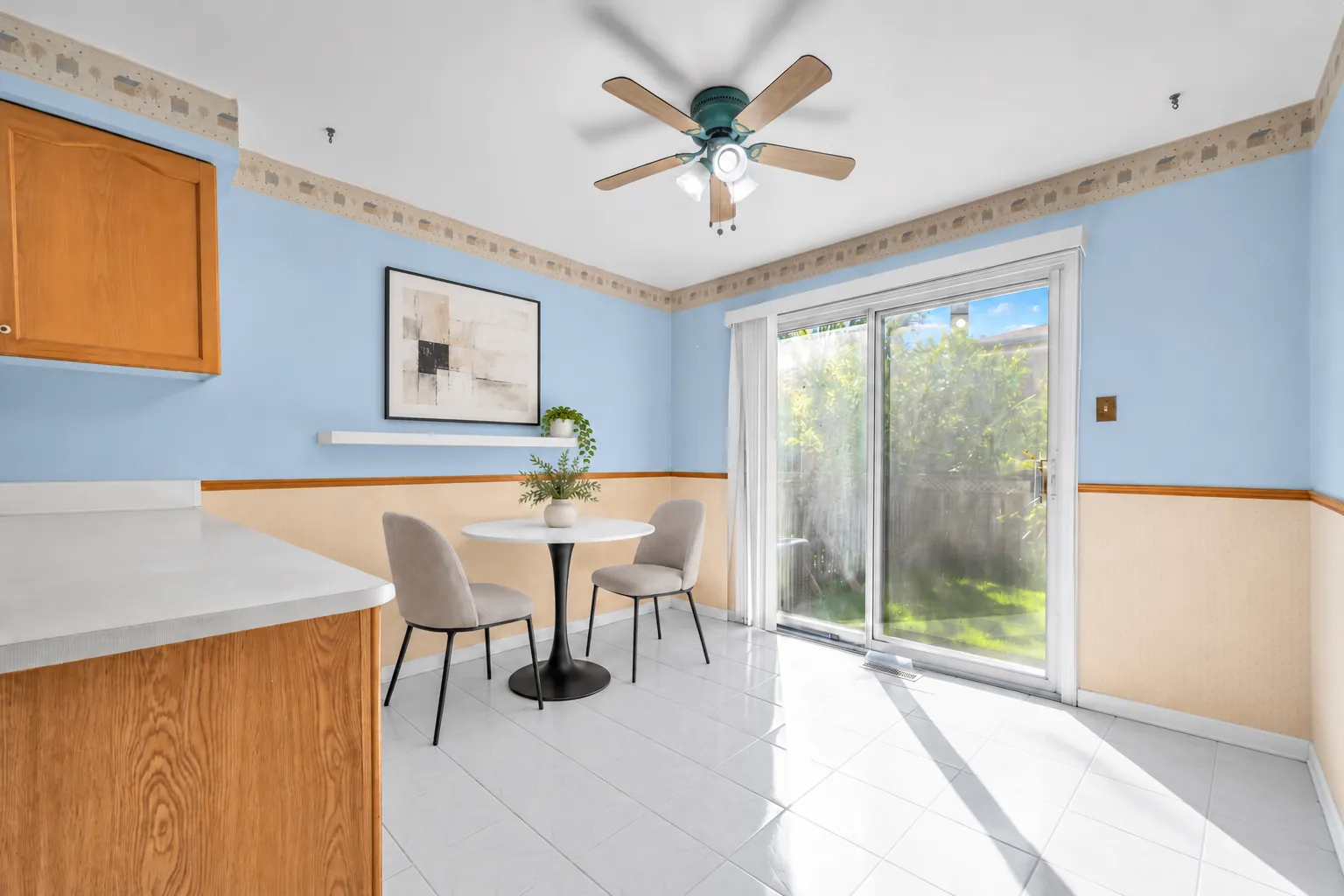 The breakfast area virtually staged: a round table and two chairs by a sliding glass door to the yard, a ceiling fan, a print on the blue wall