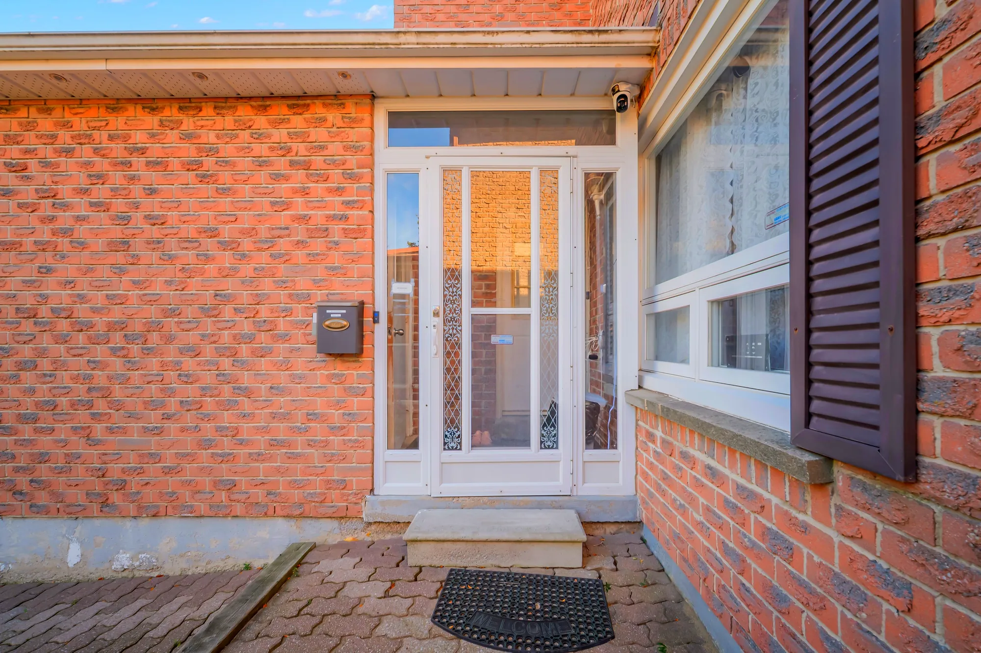 The front door: a white storm door in a brick wall, a mailbox, a window with dark shutters, a step and an interlocking walk