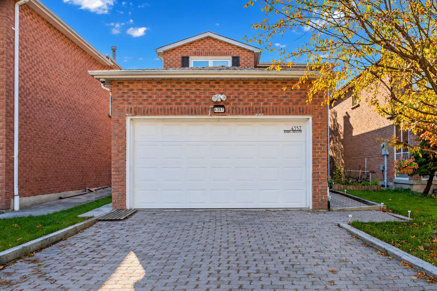 The front of 4357 Jenkins Crescent, an edited photograph: a two-storey house of red brick with a white double garage door, a long interlocking drive and a maple in autumn colour