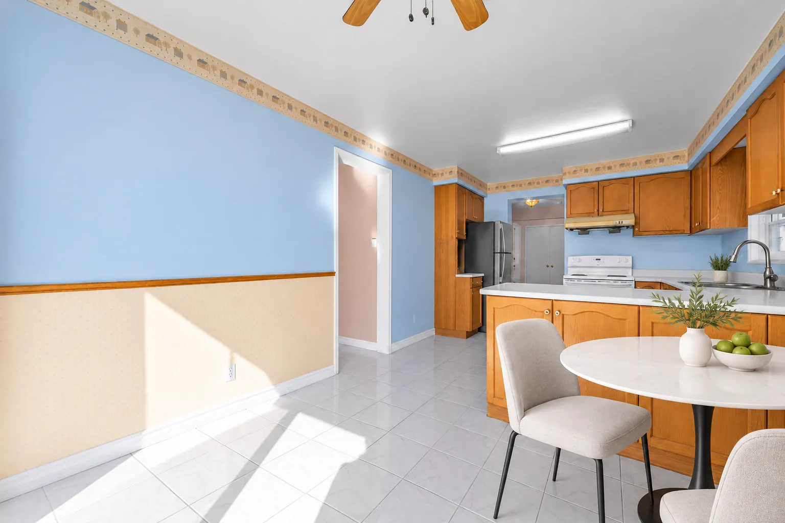 The kitchen from the breakfast area, virtually staged: the peninsula and the oak cabinets, a round table with fruit, chairs, a tiled floor in sunlight