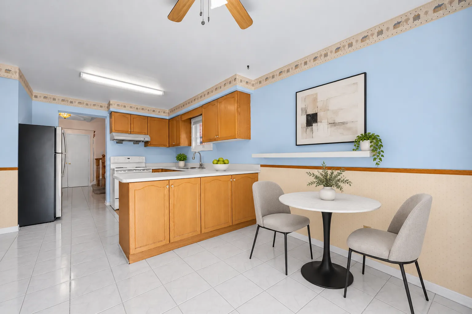 The kitchen virtually staged: oak cabinets and a peninsula, a white range, a stainless fridge, blue walls with a patterned border, a round white table and two chairs on the tiled floor