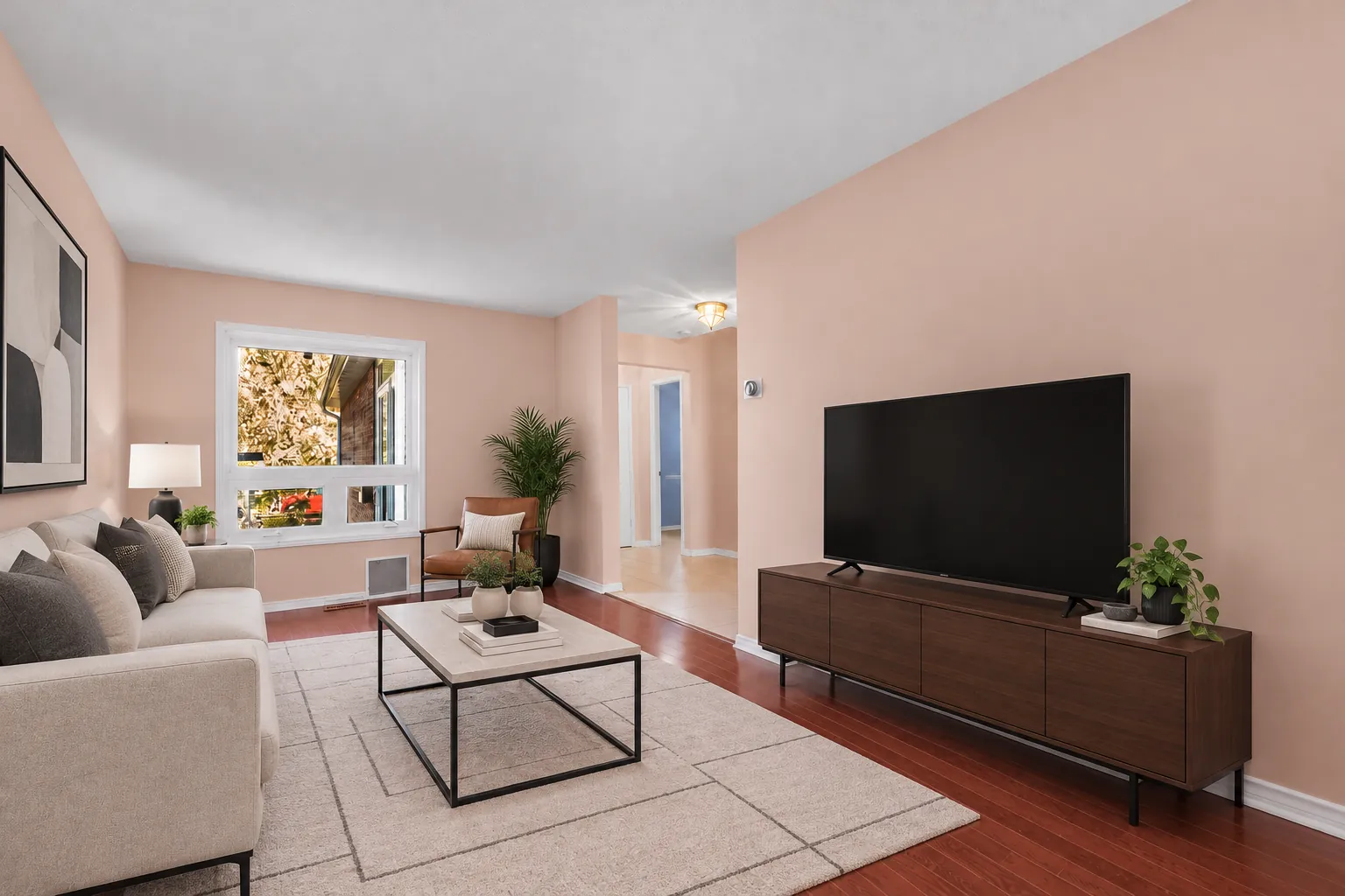 The living room virtually staged toward its window: a sofa and a lounge chair, a coffee table on a rug, a television on a low stand, the hall beyond
