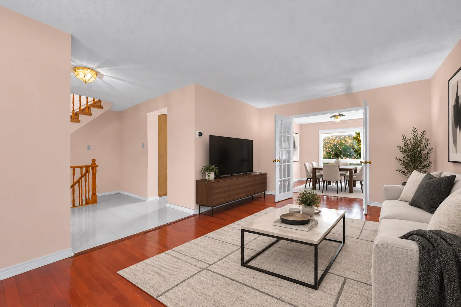 The living room virtually staged: a pale sofa, a black coffee table and a rug, a television on a wood stand, the stairs to the upper floor at the left and a pair of French doors to the dining room