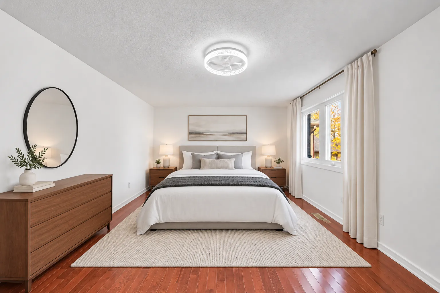 The primary bedroom from the other end, virtually staged: the bed between two night tables, a wood dresser, a round mirror, a window with long curtains