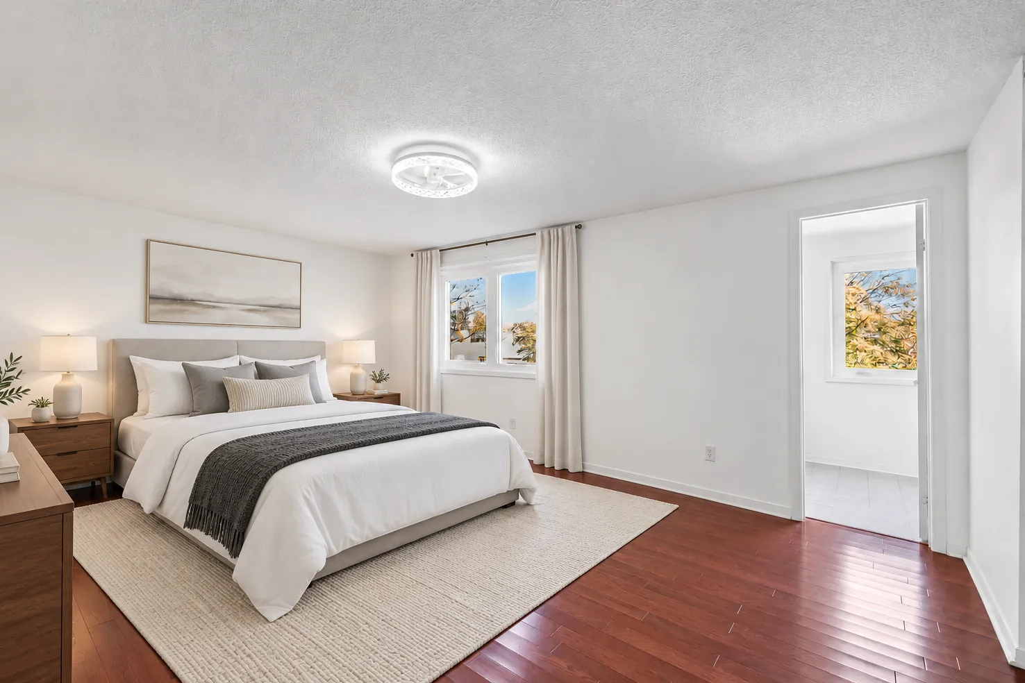 The primary bedroom virtually staged: an upholstered bed with white and grey bedding, wood night tables and lamps, a dresser, a rug, a window with curtains and a door to the bathroom